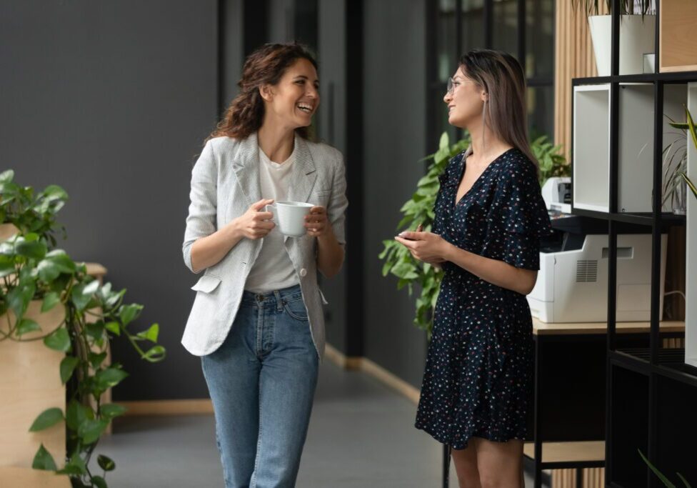 Asian and Caucasian ethnicity women colleagues met in office hall chatting enjoy friendly warm conversation, multi-ethnic mates having informal talk drink tea or coffee take break distracted from work