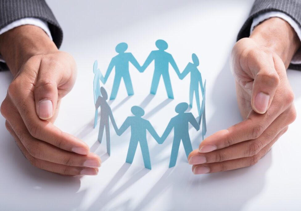 Close-up Of A Businessperson's Hand Protecting Cut-out Figures On Desk